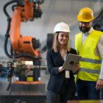 Chefingenieur und Projektleiter in einer modernen industriellen Fabrik mit Roboterarmen, die über die Optimierung der Produktion sprechen und diese planen.