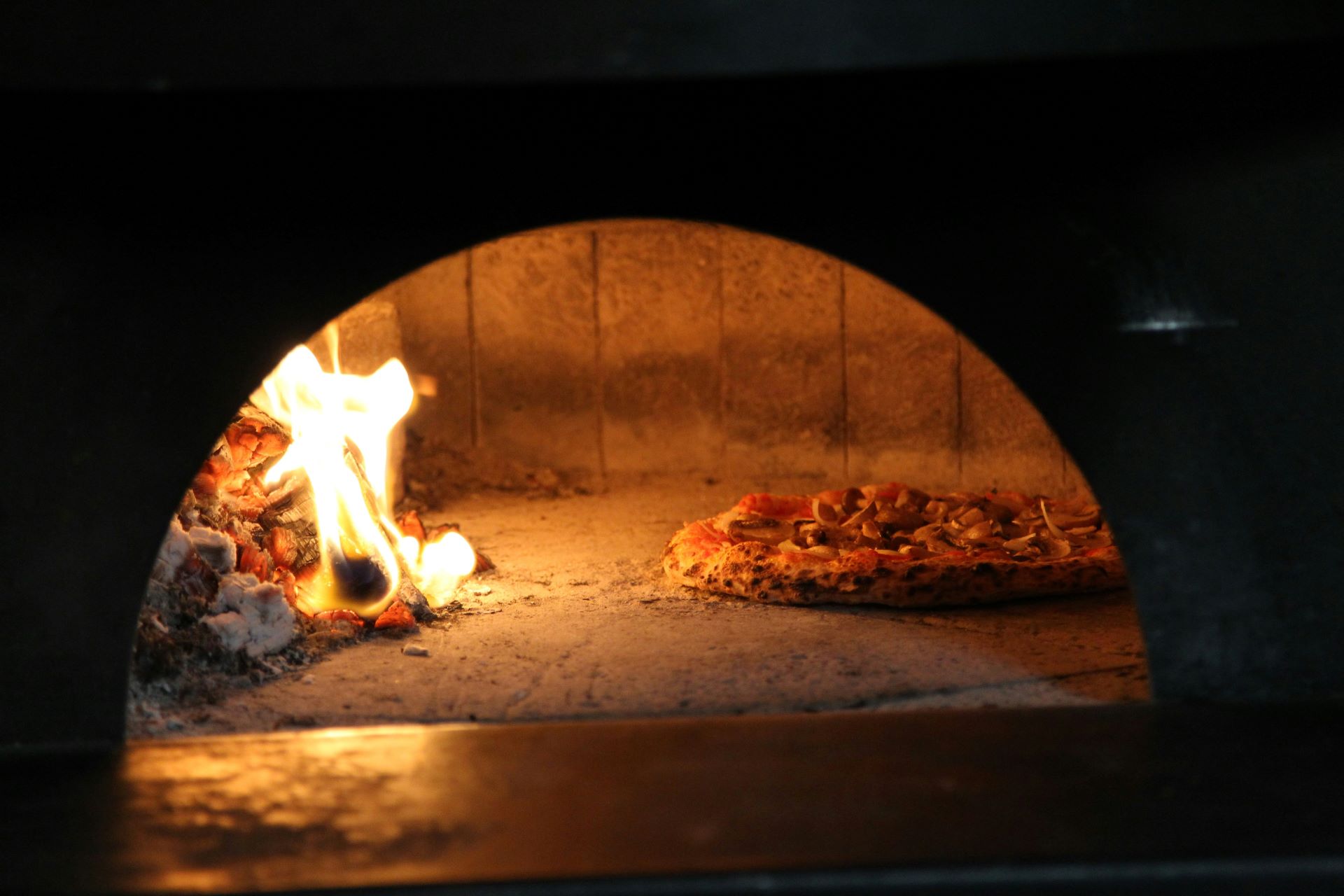 Pizza in einem traditionellen Holzofen mit glühenden Flammen und knusprigem Boden, der auf einer Steinplatte gebacken wird.