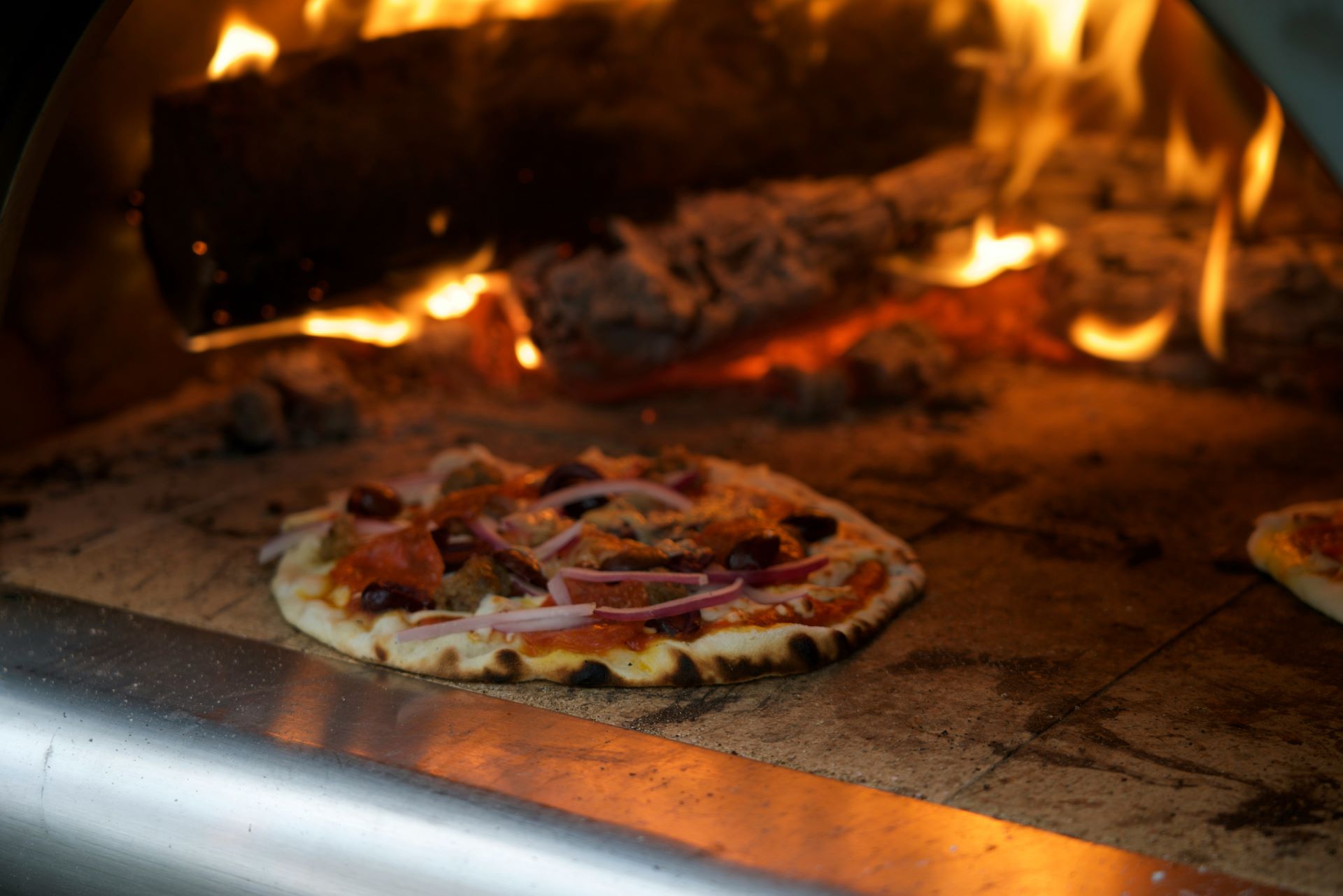 Nahaufnahme einer Pizza im Inneren eines Holzofens, umgeben von glühenden Kohlen und lebhaften Flammen.