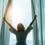 Frau in einem Hotelzimmer oder einer Ferienwohnung öffnet morgens die Vorhänge und lässt Sonnenlicht in den Raum