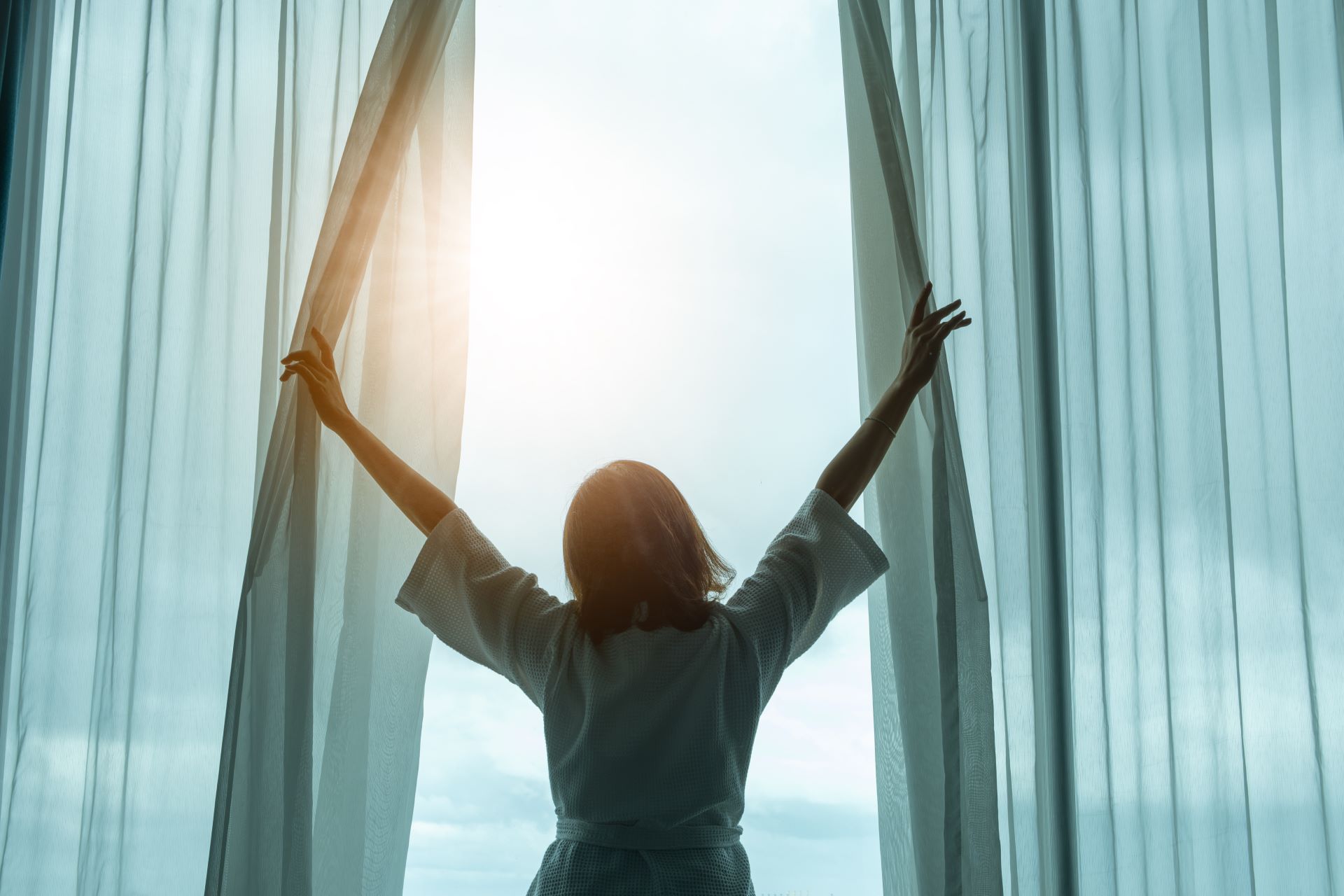 Frau in einem Hotelzimmer oder einer Ferienwohnung öffnet morgens die Vorhänge und lässt Sonnenlicht in den Raum