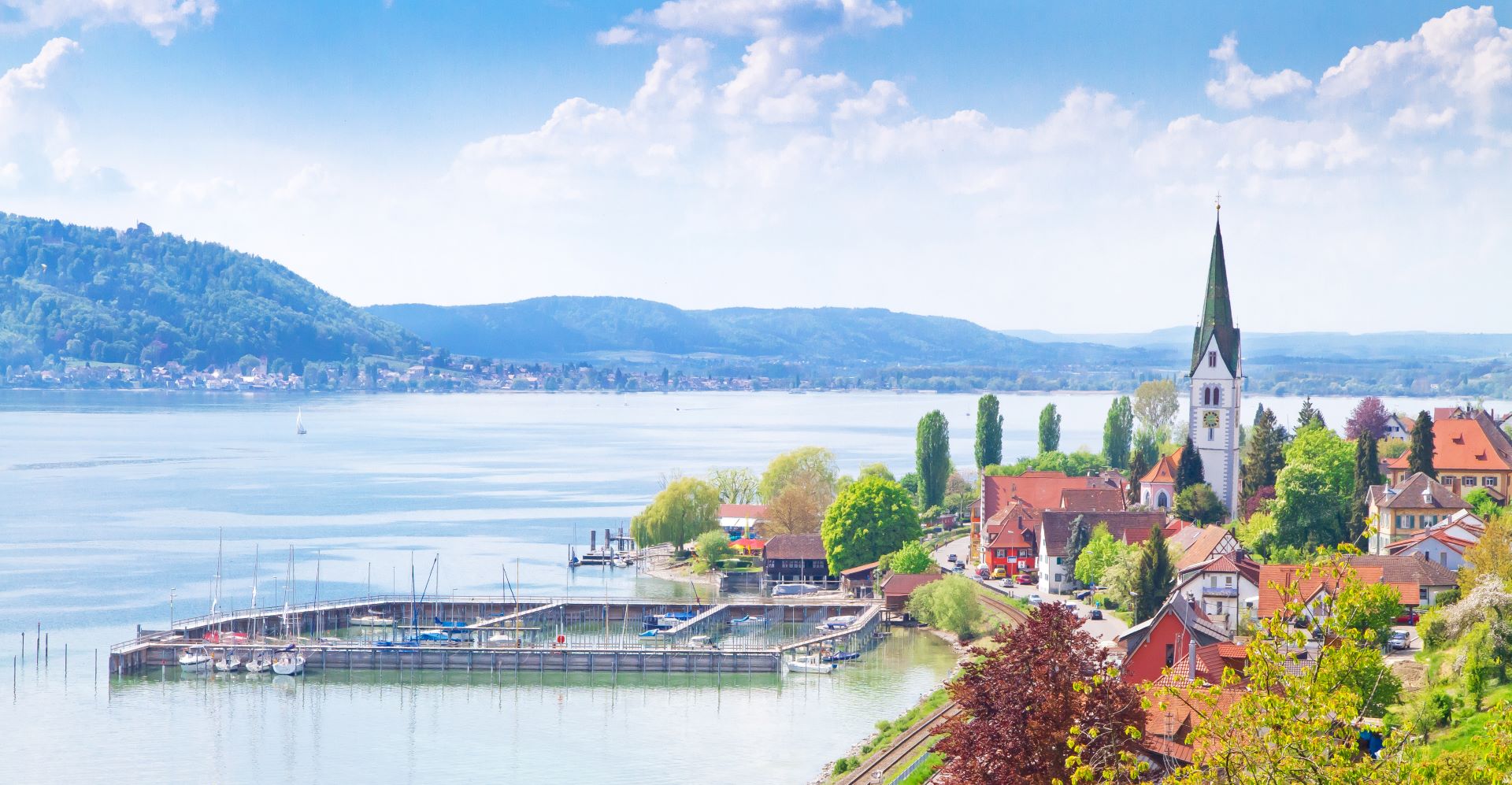 Blick auf einen malerischen Ort am Bodensee mit Hafen, Segelbooten und einer Kirche im Grünen.