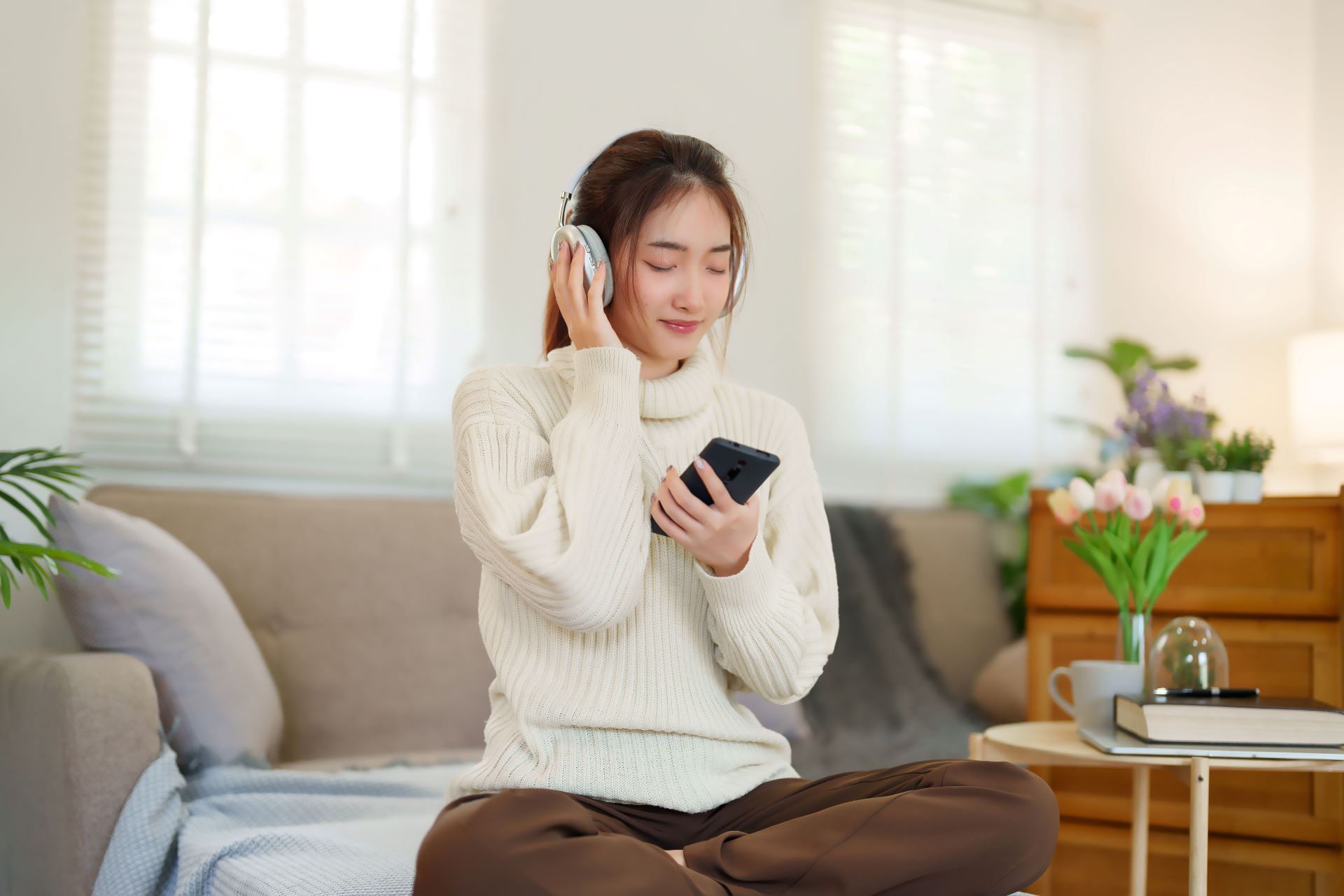 Junge Frau sitzt entspannt auf dem Sofa, hört Musik mit Kopfhörern und genießt die Atmosphäre in einer gemütlichen Ferienwohnung